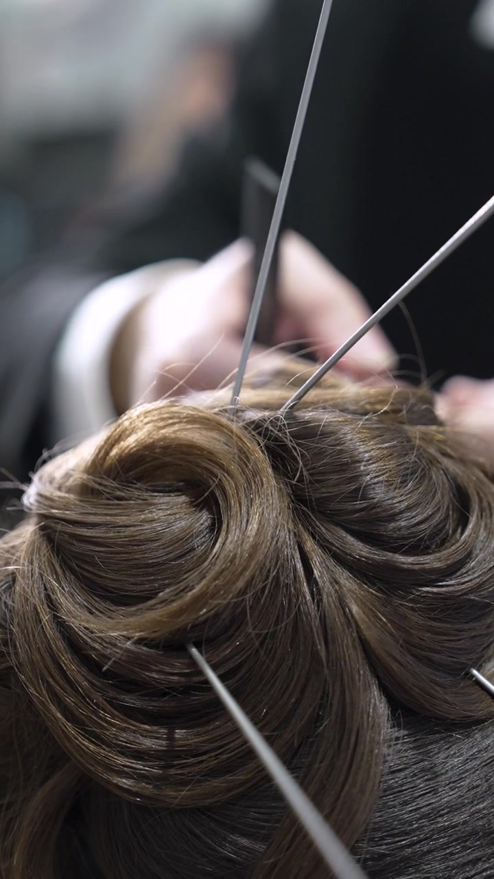 A person styles hair at a salon, focusing on an updo. Tools are used to secure the hair in place. The setting is a busy salon with various clients and stylists nearby.