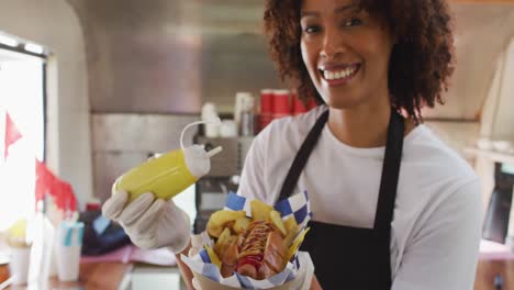retrato de una mujer afroamericana con un delantal poniendo mostaza sobre el perrito caliente en un camión de comida