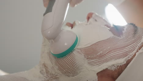 aesthetician wearing protective gloves uses vibrating electric face roller to massage jawline of spa guest applying thick white facial mask under bright lamp in clinic for smooth skin treatment