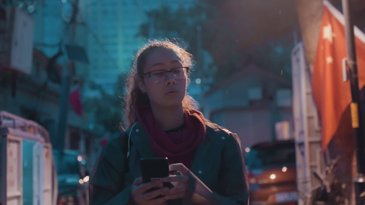 Nærbilde av kvinne som sjekker telefonen under gatelys, briller og skjerf rammer inn fokusert blikk, myke bokeh-høydepunkter, subtil følelse og håndholdt interaksjon, rolig filmatisk byatmosfære og varm lampe.