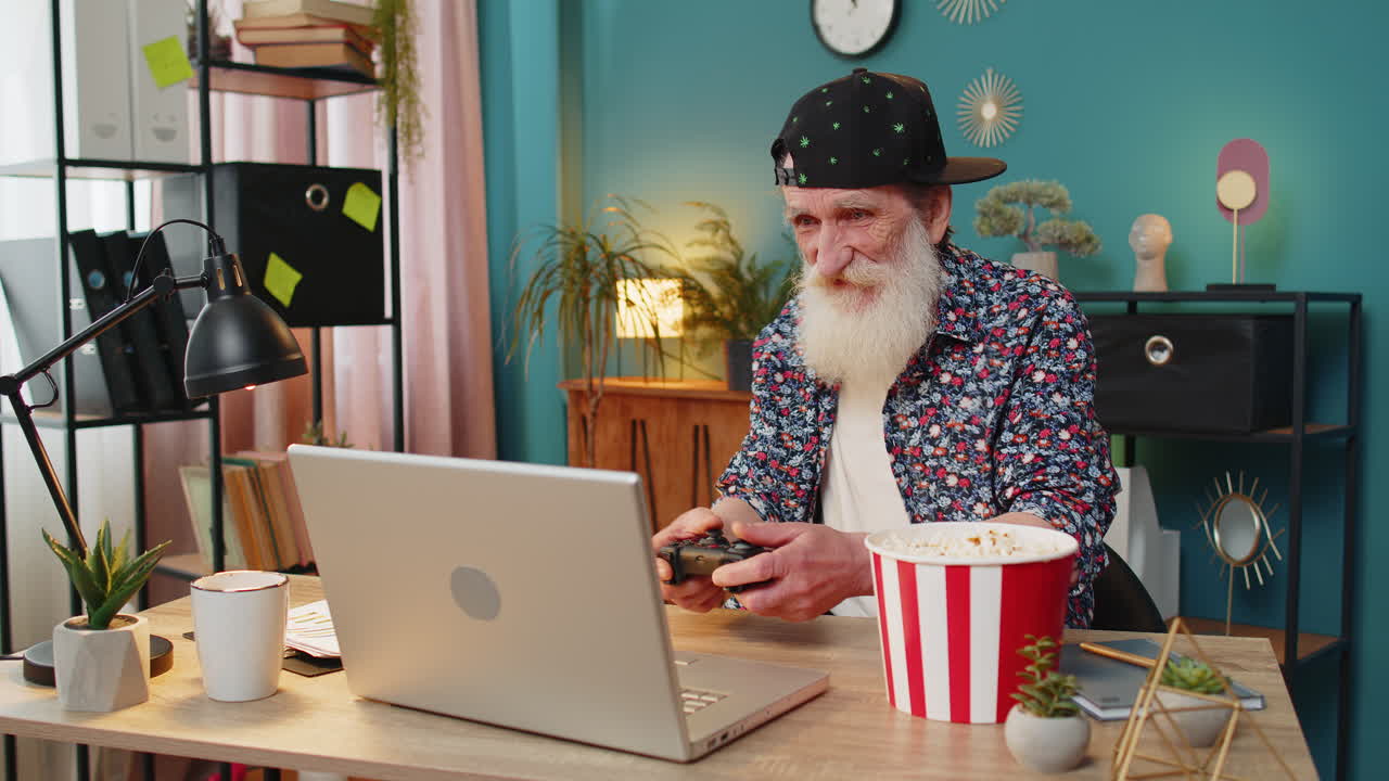Opstemt glad ældre mand, der fejrer sejr i videospil på bærbar computer ved hjælp af controller-joystick
