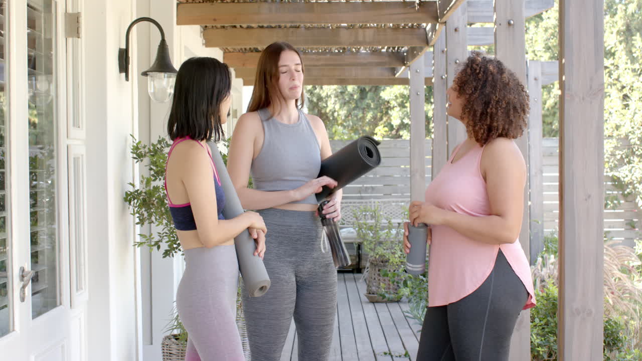 Holding yoga mats, three women friends chatting and preparing for outdoor workout