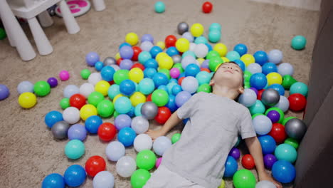 caucasian toddler lies down on the colorful balls scattered on the floor. happy active boy has fun time at home.