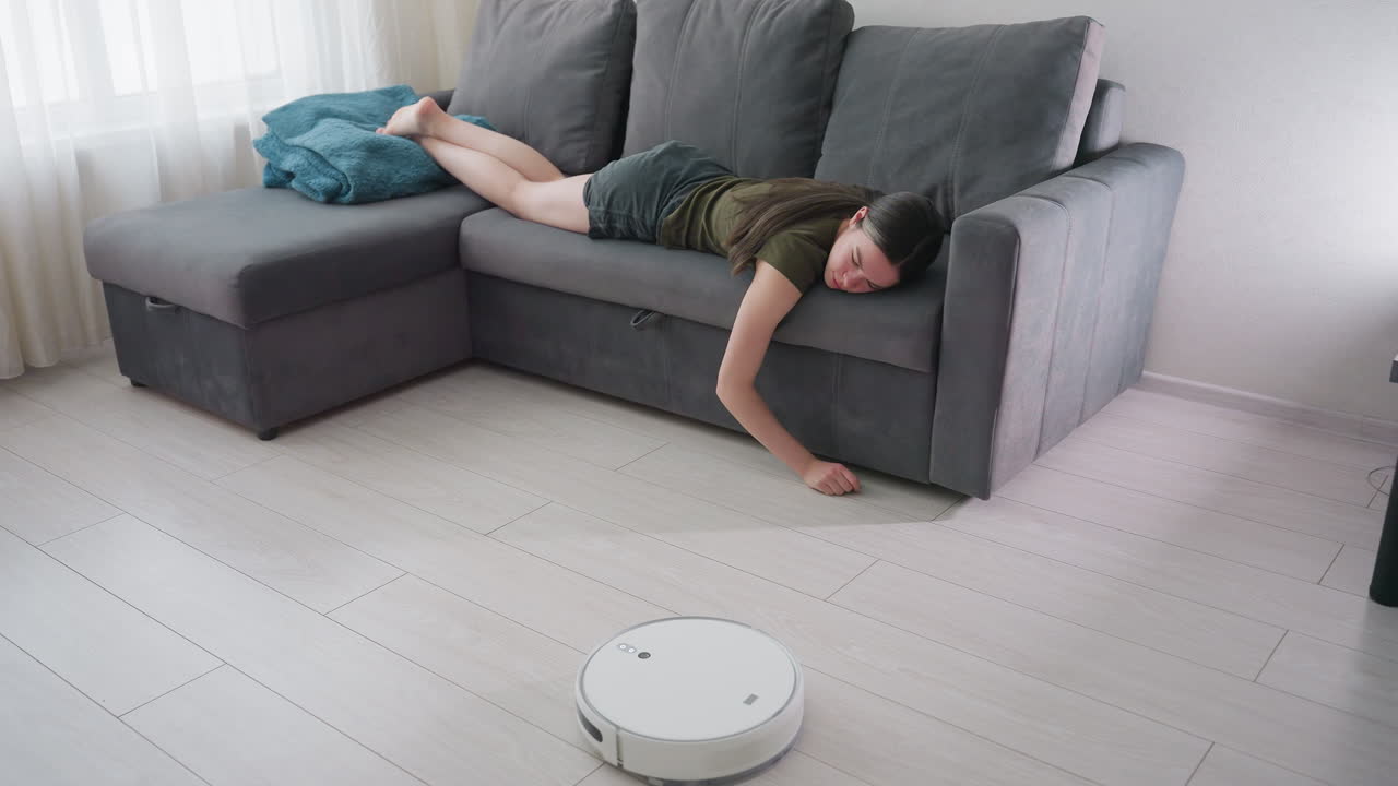 Lady lies comfortably on gray couch with hand placed on floor while robot vacuum cleaner passes by, illustrating daily life with modern home cleaning technology and relaxed household atmosphere
