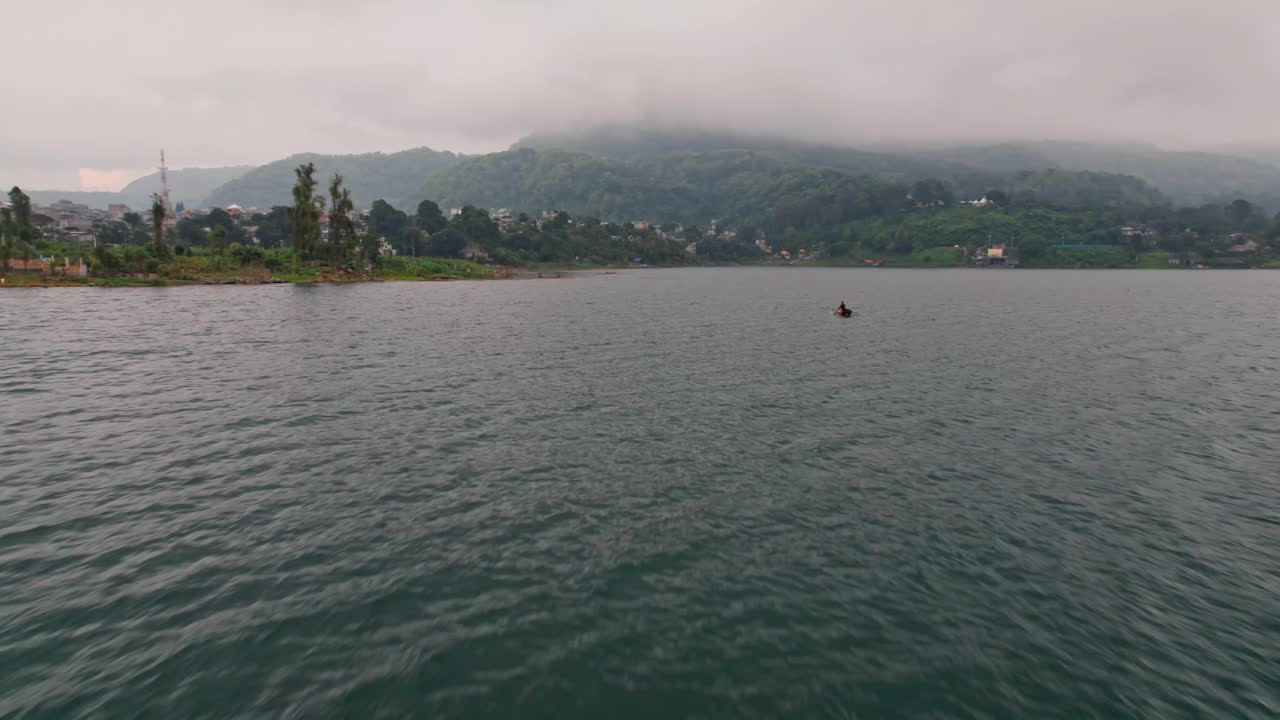 A serene drone shot captures Santiago de Atitlán Town with a lone fisherman in his traditional cayuco on the tranquil waters of Lake Atitlán at sunrise