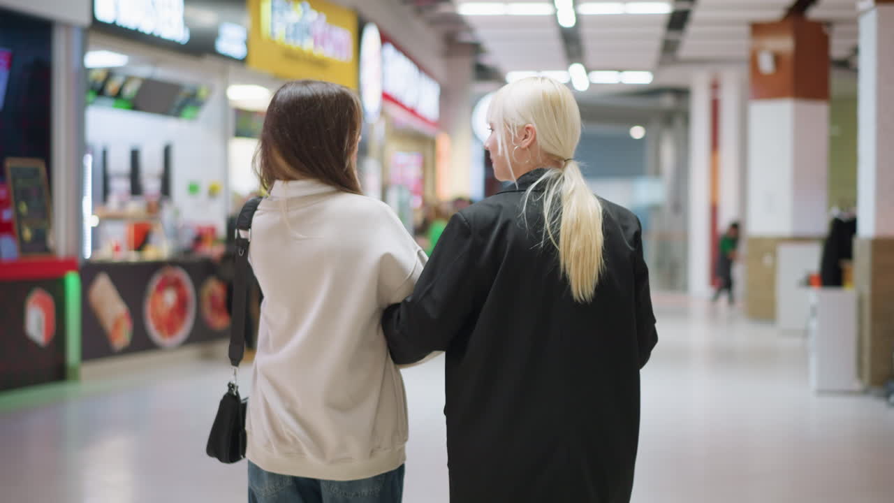 Back view of two sisters walking hand in hand through modern mall, sharing conversation, enjoying leisure time, bonding, supporting each other, exploring shops and representing friendship