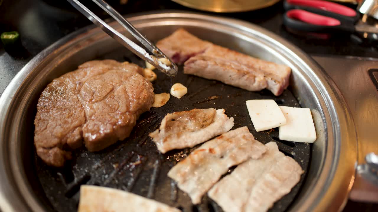 Hand uses tongs to turn garlic on tabletop Korean BBQ grill with meat and vegetables