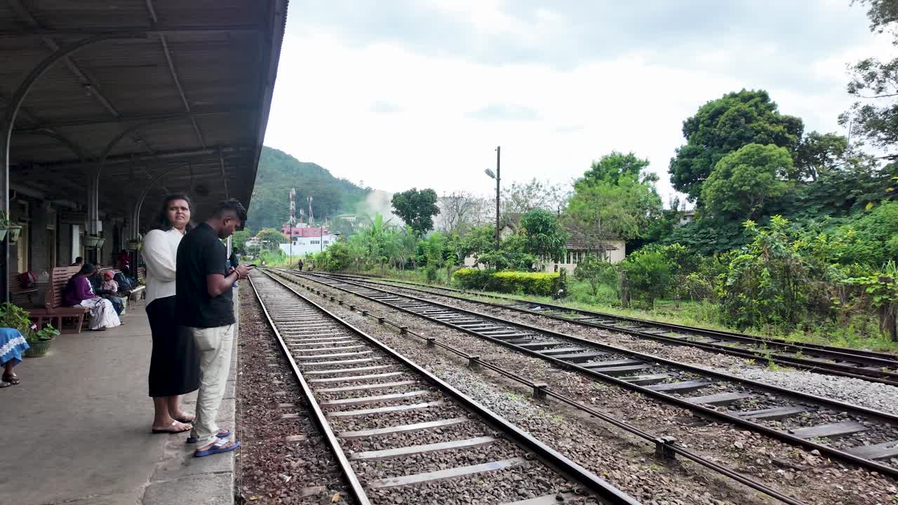 A couple waits patiently at the picturesque Haputale railway station in Sri Lanka, taking in the serene surroundings and lush greenery while checking their mobile phones.