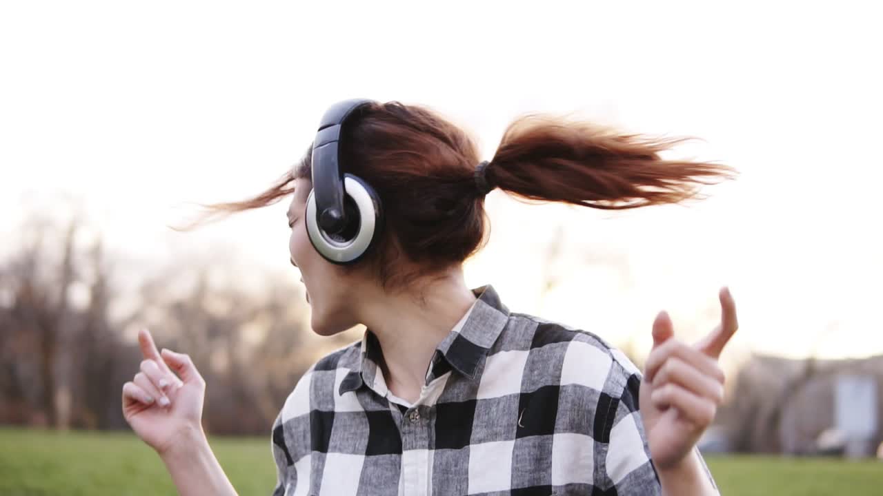 A girl with brown hair is very emotionally and expressively waving her head listening to music on headphones. Dancing. Gesturing with both hands. Portrait. Outside in the park
