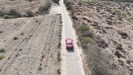 Toma-Aérea-Reveladora-De-Un-Vehículo-Rojo-En-Movimiento-A-Lo-Largo-De-Una-Carretera-Polvorienta-Y-Montañosa-En-El-Desierto-En-Perú-En-El-Camino-A-La-Ciudad-Santa-De-Caral-Con-Vista-De-árboles-Marchitos-Y-Montañas-En-Un-Día-Soleado