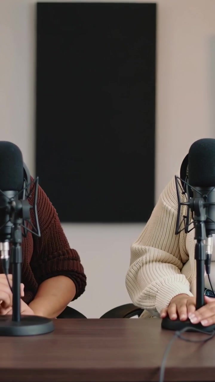 Video verticale: Due conduttori in maglione controllano appunti, chinati verso i microfoni in studio per un podcast.