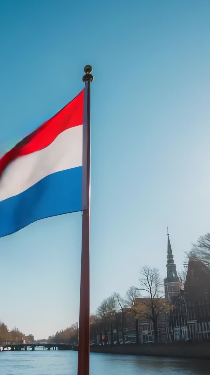 Vertical video: Fluttering Dutch flag responding to rising breeze beside canal, with church spire