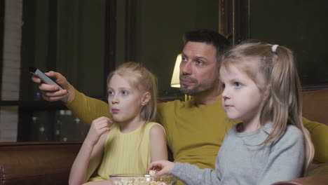 Papá-Con-Control-Remoto-De-Tv-Y-Sus-Dos-Hijas-Pequeñas-Comiendo-Palomitas-De-Maíz-Y-Viendo-Una-Película-Mientras-Se-Sientan-En-El-Sofá-Por-La-Noche-En-Casa