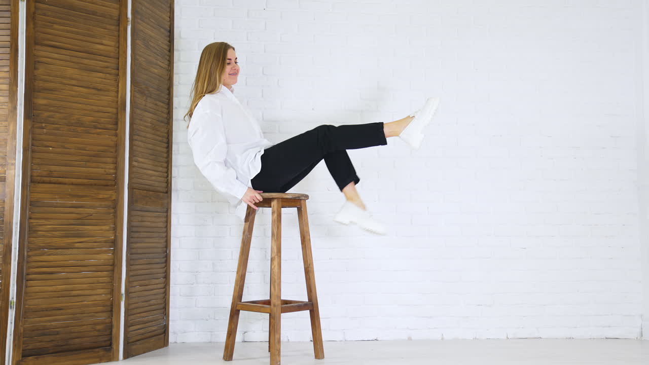 Smiling lady sitting on high stool and waving legs. Demonstrating new white shoes at the backdrop of white wall and wooden doors.