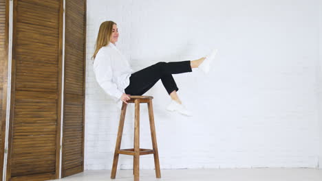 smiling lady sitting on high stool and waving legs. demonstrating new white shoes at the backdrop of white wall and wooden doors.