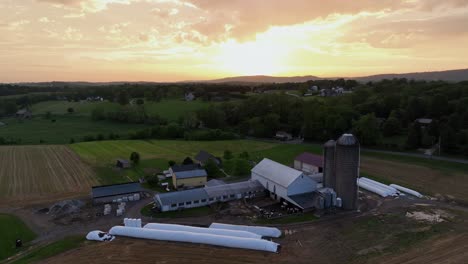 American farm with covered hay bales, green house, stable, barn , silo storage and farmers house. Aerial rising wide shot. Golden sunrise behind farm fields in rural district of american town.