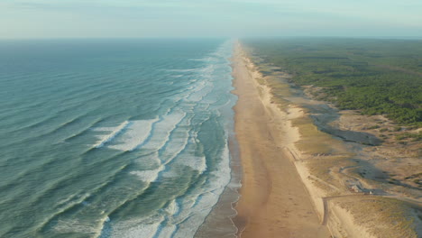 Schöner-Leerer-Strand-Zur-Goldenen-Stunde,-Der-Endlos-In-Die-Ferne-Mit-Grünen-Wäldern-Und-Blauem-Ozean-Reicht