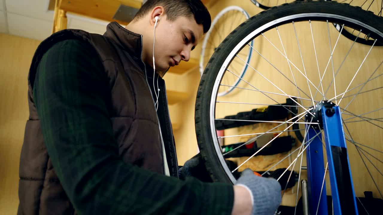 el mecánico concentrado está fijando los radios en la rueda de la bicicleta con herramientas especiales mientras hace el servicio de la bicicleta y escucha música a través de auriculares. mantenimiento y concepto de profesión.
