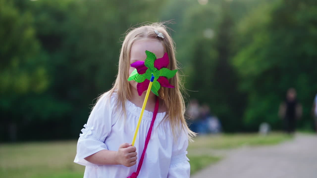 Beautiful little girl with colorful toy in the park. Pretty young child in white blouse looking at camera joyfully.