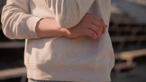 close-up of woman s hands folded in front, wearing a soft, casual hoodie with warm sunlight gently highlighting her arms, the background is softly blurred featuring empty bleachers