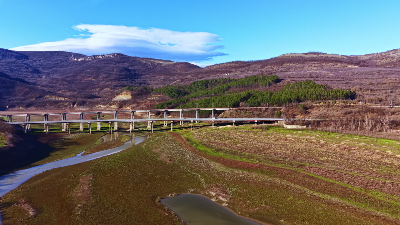 View of a bridge over a river in a valley surrounded by hills during a clear day with few clouds in the sky and dry vegetation on the ground