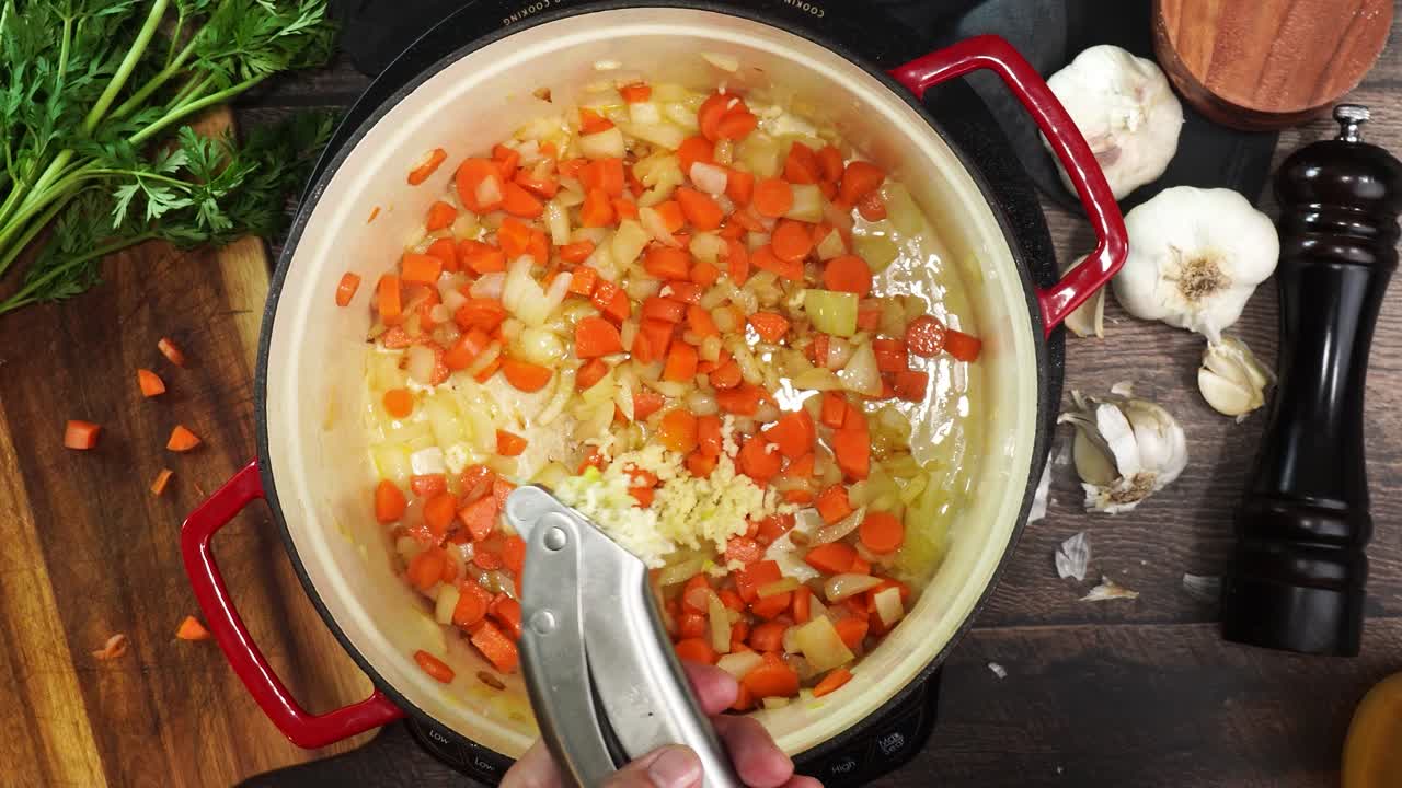 Cooking Italian Wedding Soup and adding in minced garlic using a garlic press to the red stock pot. Using a wooden spoon to stir the soup while its simmering. Adding garlic while cooking soup.
