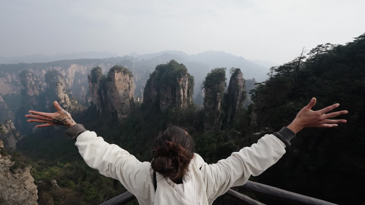 Person opens arms wide facing towering sandstone pillars and misty mountain ridges of Zhangjiajie, captured in a sweeping scenic moment