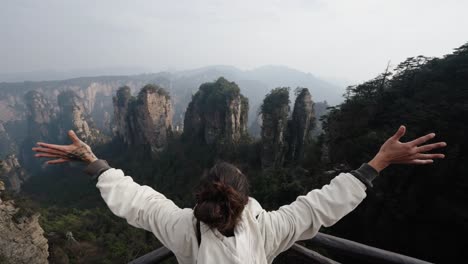 person opens arms wide facing towering sandstone pillars and misty mountain ridges of zhangjiajie, captured in a sweeping scenic moment