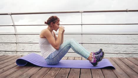 Vista-De-Cerca-De-Una-Joven-Atlética-Haciendo-Sentadillas-En-La-Alfombra-Junto-Al-Océano-Temprano-En-La-Mañana.-Mujer-Fitness-Haciendo-Abdominales