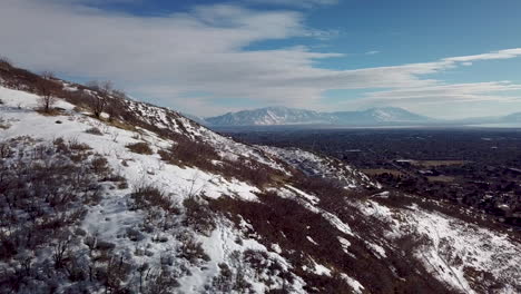 Volando-Sobre-Las-Colinas-Para-Revelar-Una-Ciudad,-Un-Lago-Y-Montañas-Al-Otro-Lado-Del-Valle-En-Invierno