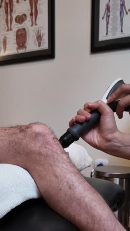 close-up of radial shockwave therapy device targeting the patellar tendon of the knee during a pain management session in a clinical rehab setting. anatomical chart and equipment in the background.