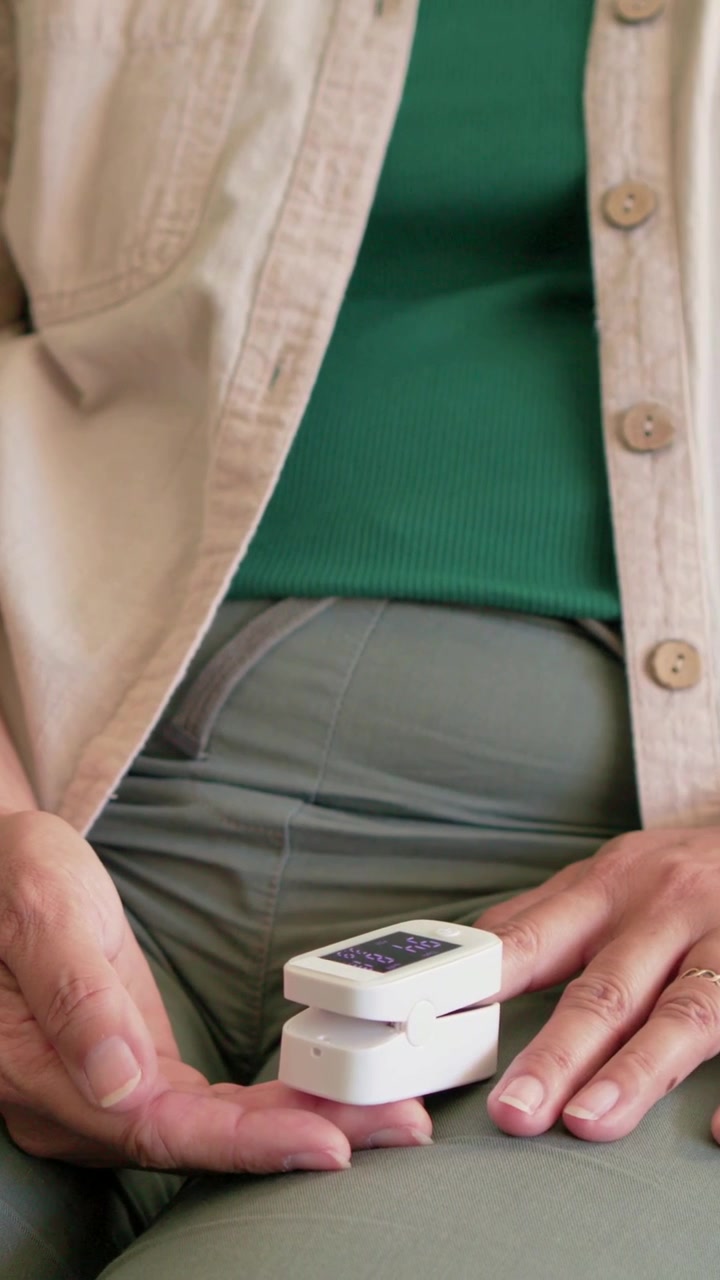 Vertical video: Moving hand, woman inserting finger into oximeter to measure SpO2 on lap, green top