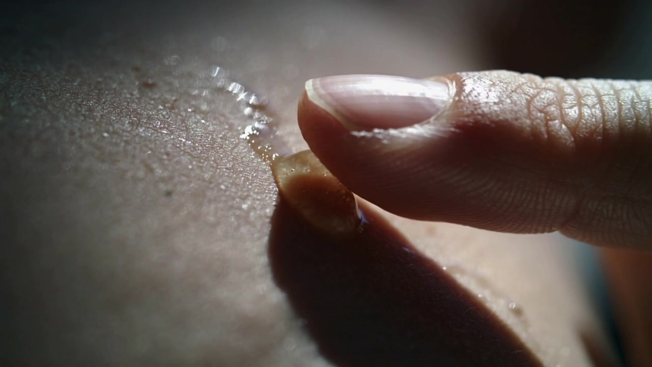 Polpastrello che si avvicina e tocca una goccia ambrata sulla pelle nuda, premendo e spalmando il liquido per stenderlo in uno strato sottile.