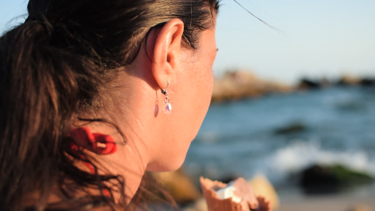 captura de pantalla de una hermosa mujer con un aspecto natural comiendo helado en la playa mientras las olas rompen y el sol ilumina su rostro