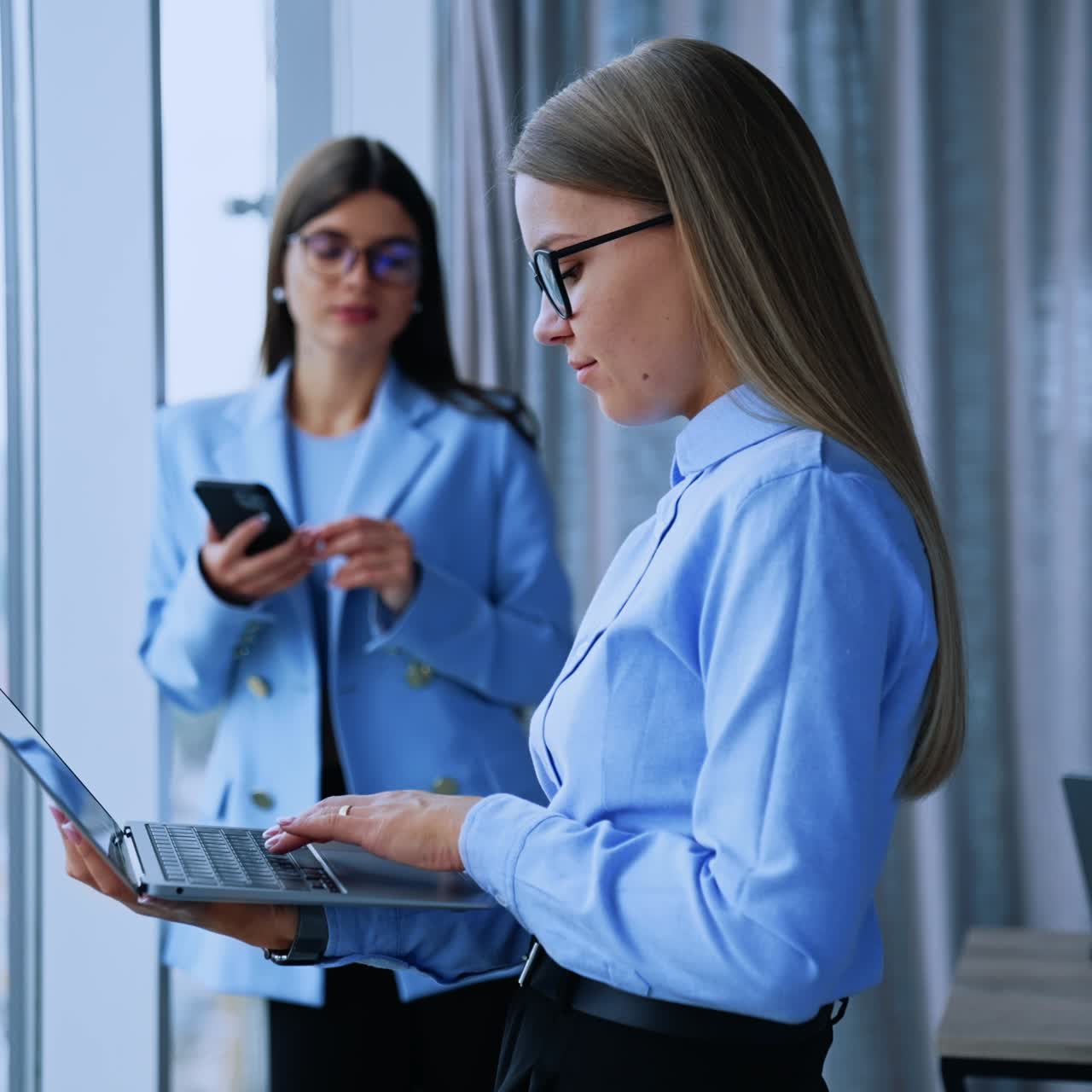 Using modern technologies at work. Young ladies stand in office holding laptop and smartphone. Blurred backdrop