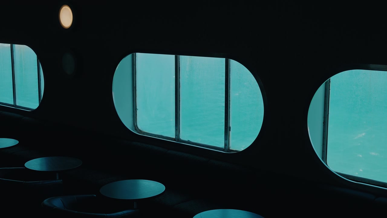 Grandi finestrini arrotondati di un traghetto che brillano di blu accanto a tavoli vuoti in una cabina passeggeri fiocamente illuminata.