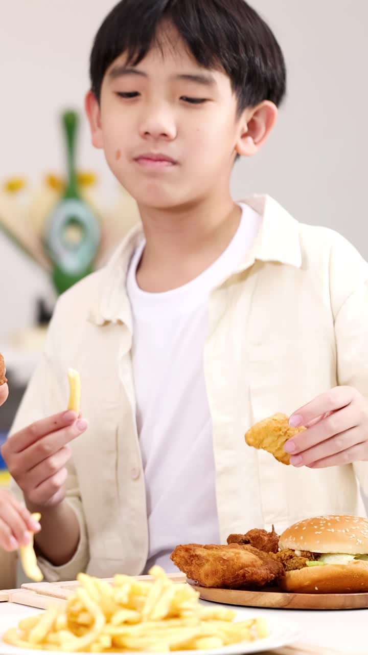 明るい教室のテーブルでフライドチキンとフライドポテトを食べる笑顔のアジア人少年