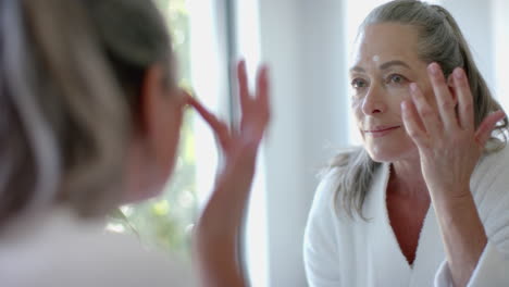 applying skincare, mature woman in bathrobe looking in mirror at home, smiling