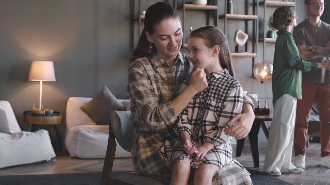 Happy Mom With Her Little Girl Sitting On Chair At Home And Smiling Together At Camera While In The Background Family Talking To Each Other