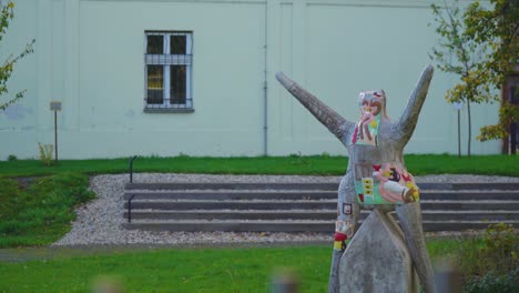 weird looking artistic statue made out of wood with arms extended in the air in the middle of a garden behind a fence steps in the background romance building czech republic opava