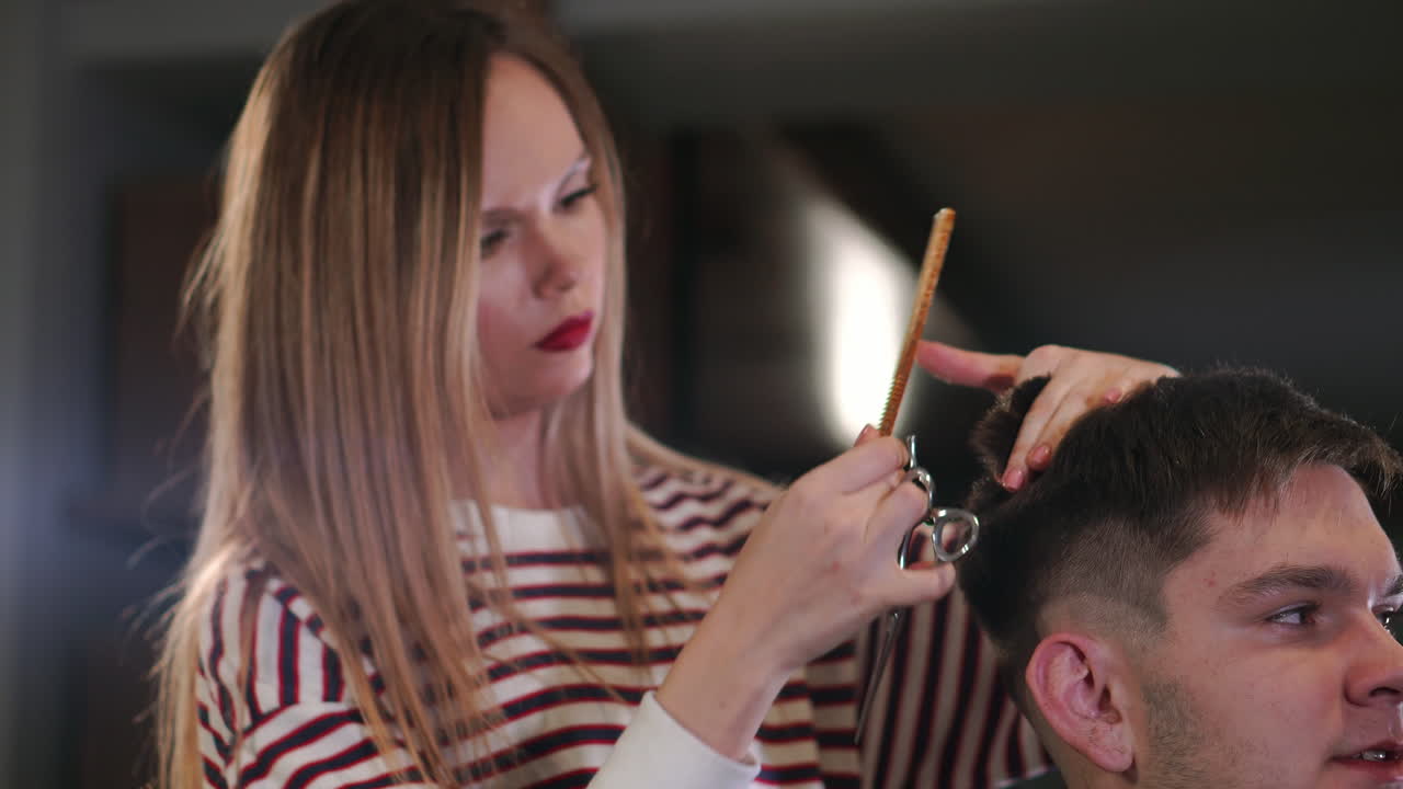 Interior shot of working process in modern barbershop. Side view portrait of attractive young man getting trendy haircut. Male hairdresser serving client, making haircut using metal scissors and comb.