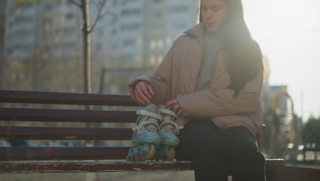 close-up of a young girl wearing a peach jacket, grey inner shirt, and black trousers walking through a sunlit park. she sits on a bench and places her rollerblades beside her