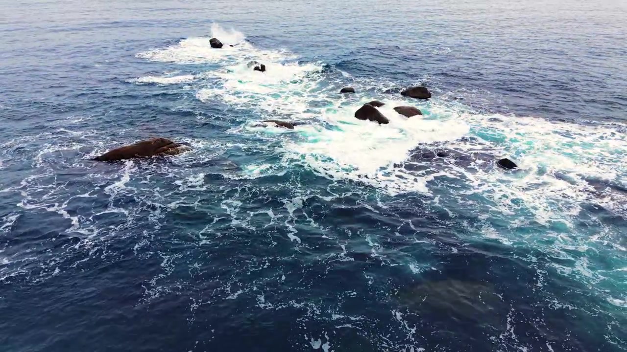 Pemandangan sudut tinggi batu-batu gelap yang tersebar di tengah buih laut putih yang berputar dan riak-riak di permukaan laut biru. Batu-batu besar pesisir yang menonjol dari air laut yang bergejolak.