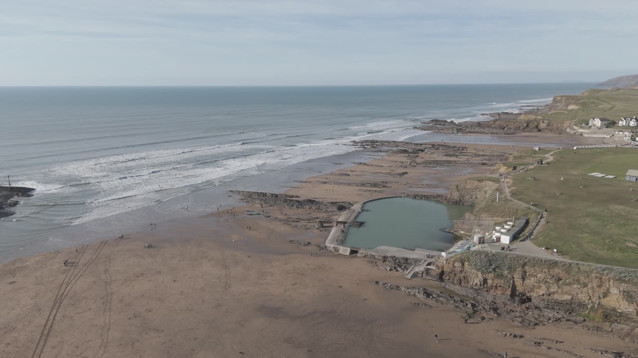 Aerial sweep of concrete-walled ocean pool beside expansive sandy beach with walkers and surfers enjoying calm tidal water against open Atlantic swells