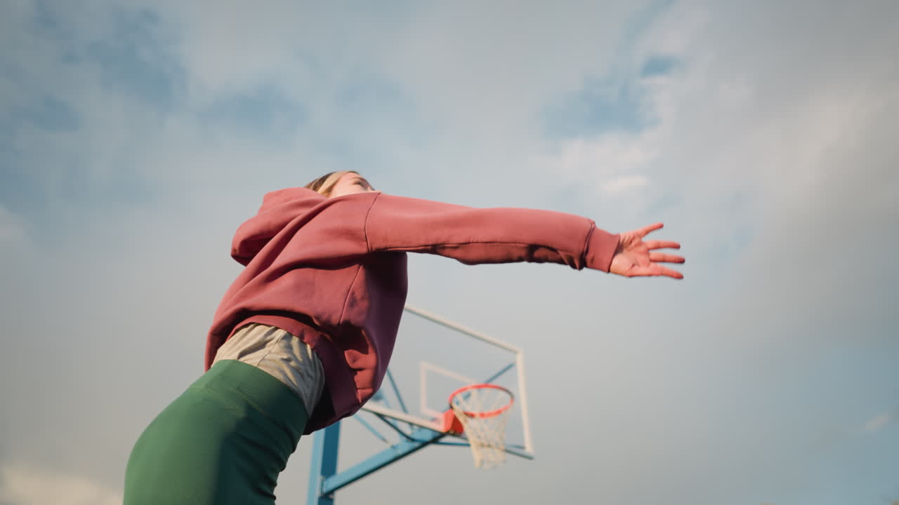 dama en ropa atlética salta alto, golpeando voleibol en la cancha al aire libre, aro de baloncesto visible en el fondo, con árboles verdes, edificios