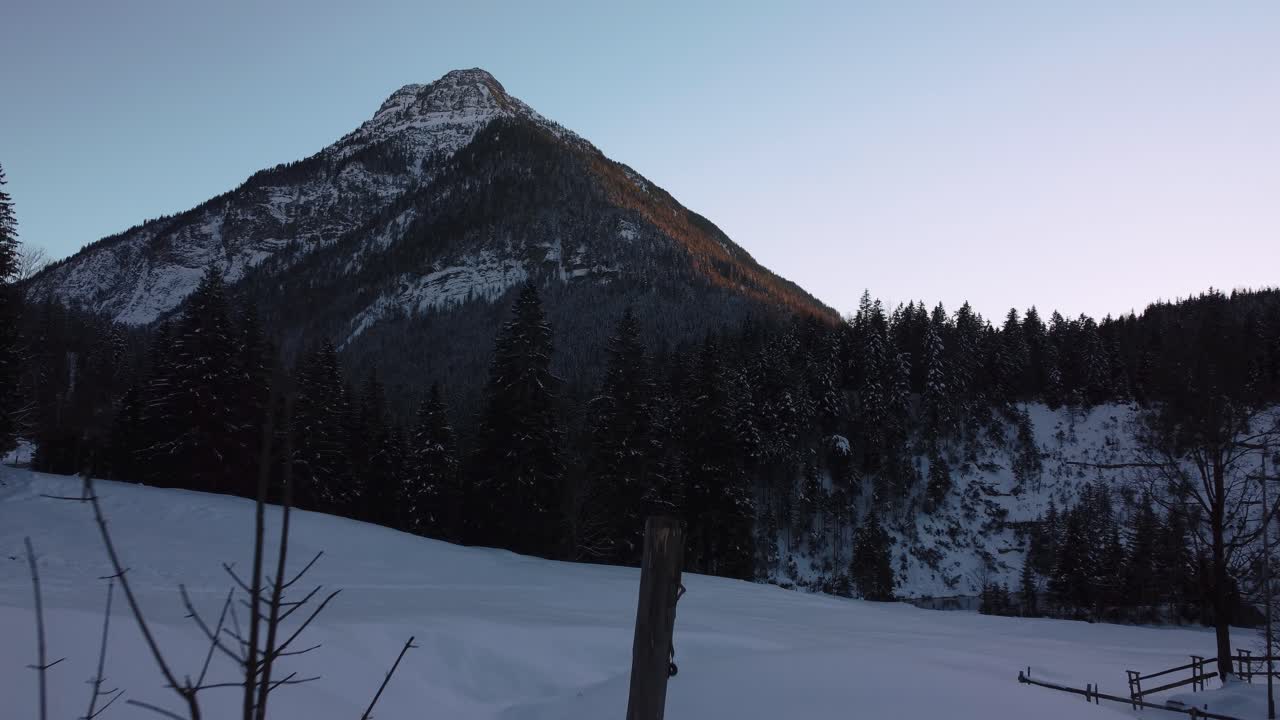 vuelo panorámico sobre la nieve hacia una enorme e impresionante montaña con bosques y árboles en invierno con nieve y hielo en las idílicas montañas bávaras de los alpes austríacos con picos rojos al atardecer