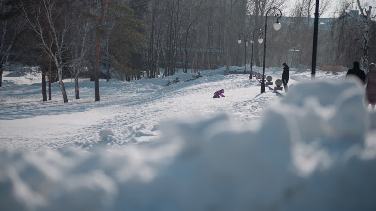 blurred foreground of fresh snowdrifts in park path with distant child playing in colorful snowsuit and adult watching under decorative lamp post bulbs beside bare trees in soft winter daylight