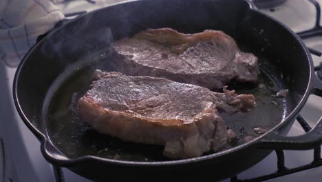 smoking steaks being halfway cooked in a cast iron skillet with oil - slow motion