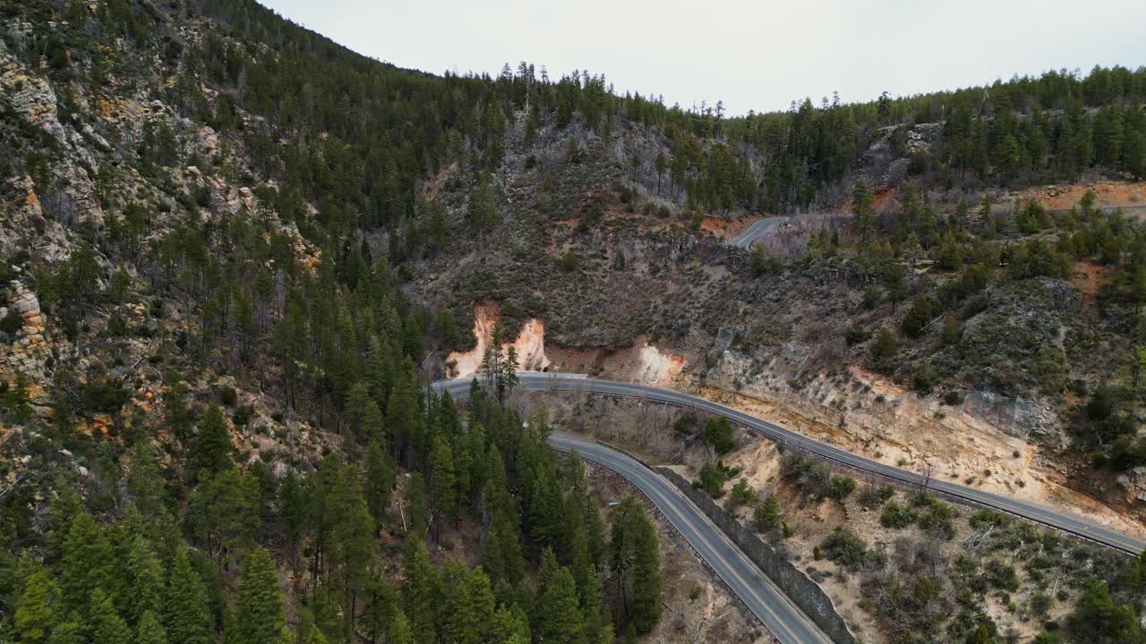 An aerial drone shot reveals winding mountain roads snaking through a dense pine forest on a cloudy day, showcasing rugged terrain and natural beauty