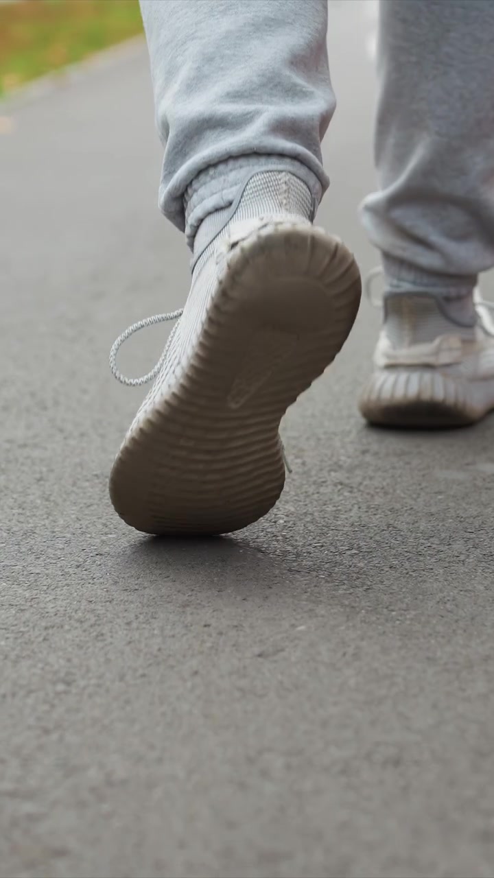 White sneakered feet move through outdoor pavement area. Worn soles show texture as shoes make contact with ground. Outdoor setting with blurred greenery behind walking path